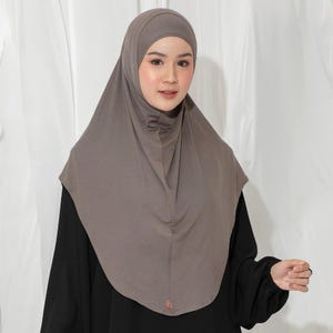 May include: A taupe-colored hijab, a head covering worn by some Muslim women. The hijab is made of a soft, draping fabric and covers the head and neck. The person in the image is wearing a black long-sleeved shirt.
