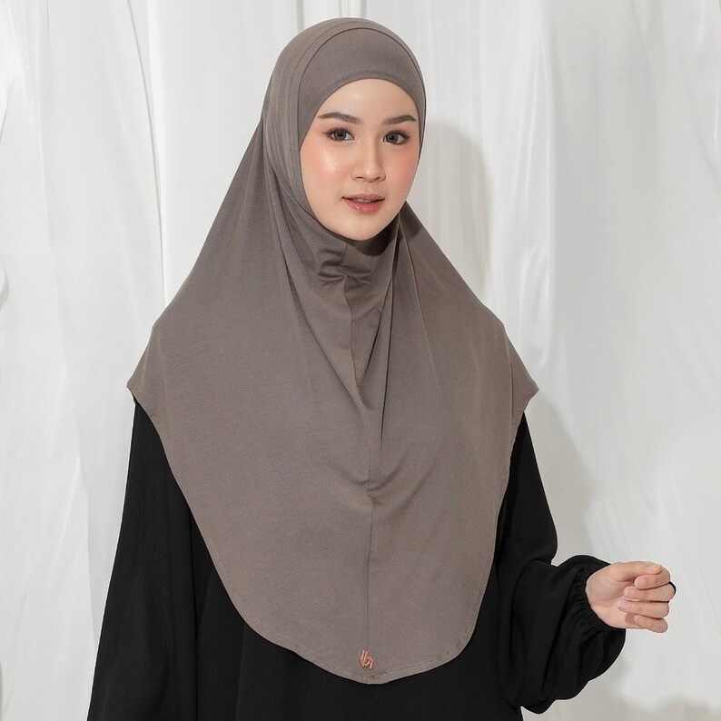 May include: A taupe-colored hijab, a head covering worn by some Muslim women. The hijab is made of a soft, draping fabric and covers the head and neck. The person in the image is wearing a black long-sleeved shirt.