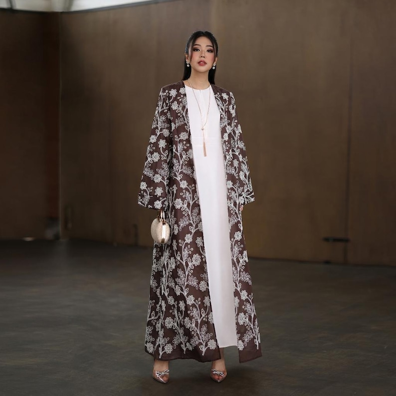 May include: A floor-length white dress paired with a long, open-front brown floral patterned coat. The coat features a white floral design. The outfit is accessorized with a gold-colored purse and a long necklace.