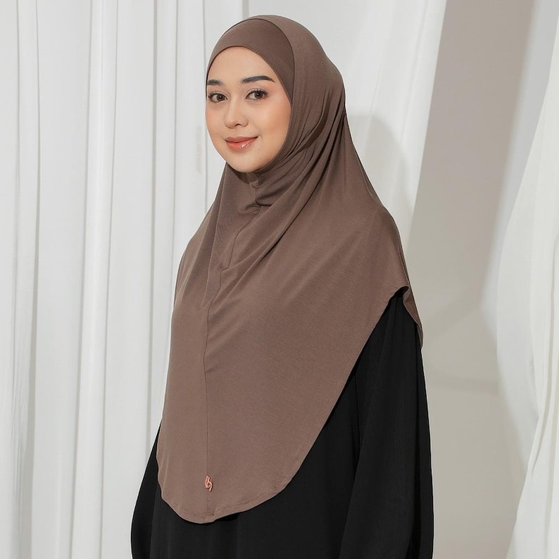 May include: A brown hijab, a head covering worn by some Muslim women, is draped over the head and shoulders. The hijab is made of a soft, flowing fabric. A small, decorative pin is visible on the front of the hijab.