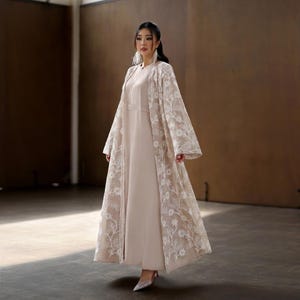 May include: A woman wearing a beige dress with a long, open, cream-colored lace overlay. The lace features a floral pattern. The dress has a modest neckline and long sleeves. The woman is wearing matching heels.