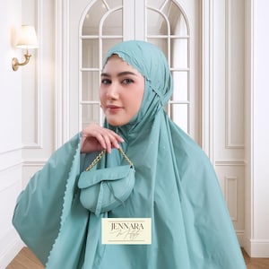 May include: A light teal prayer garment set, including a head covering and a matching small purse with a gold chain strap. The head covering has a scalloped edge and is adorned with small, sparkling embellishments. The purse is held in the hand.
