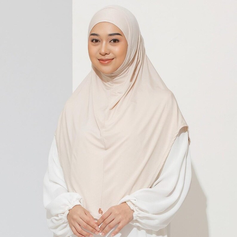 May include: A beige hijab, a head covering worn by some Muslim women, is draped over the head and shoulders. The hijab is made of a soft, flowing fabric. The person is wearing a white long-sleeved shirt underneath the hijab.