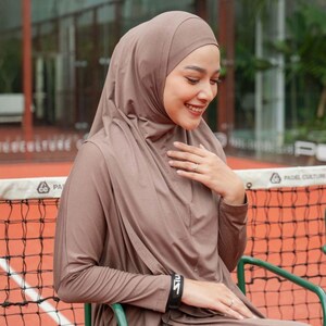 May include: A woman wearing a taupe-colored hijab and matching long-sleeved top. The outfit includes a black wristband with white text. She is seated outdoors, smiling, with a tennis net visible in the background.
