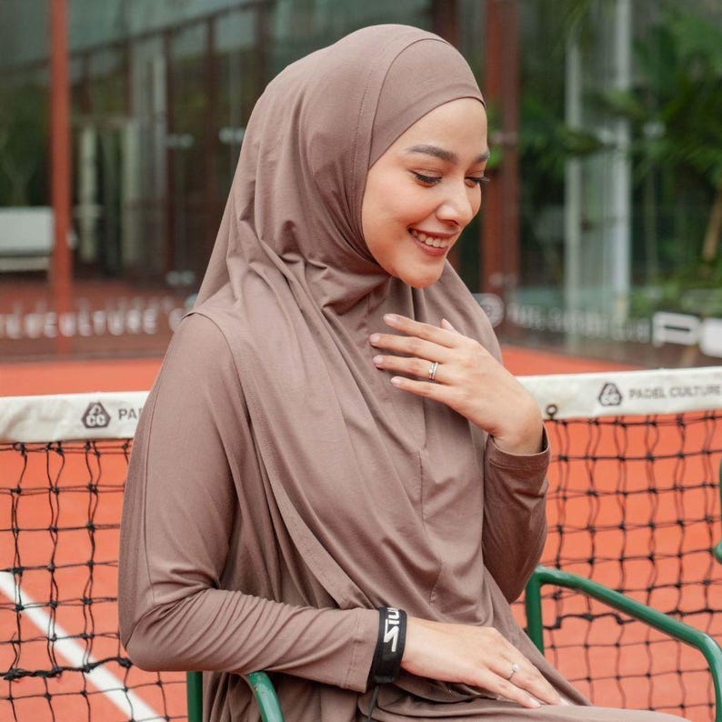 May include: A woman wearing a taupe-colored hijab and matching long-sleeved top. The outfit includes a black wristband with white text. She is seated outdoors, smiling, with a tennis net visible in the background.