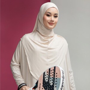 May include: A woman wearing a beige hijab and long-sleeved dress holds a padel racket. The racket has a black and white grip and a pink, white, and black face. The background is a combination of pink and gray.