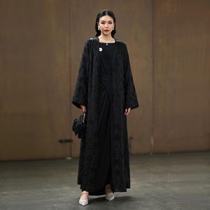 May include: A long black abaya with detailed floral patterns, paired with a black dress. The outfit is completed with a small black handbag and silver heels. A brooch and necklace provide delicate embellishments.