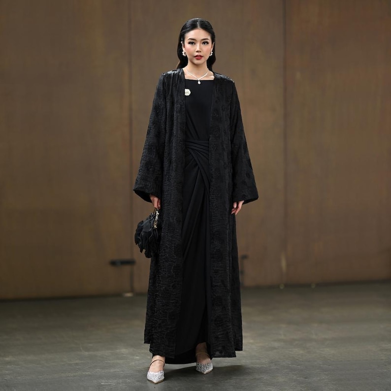 May include: A long black abaya with detailed floral patterns, paired with a black dress. The outfit is completed with a small black handbag and silver heels. A brooch and necklace provide delicate embellishments.