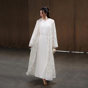 May include: A woman wearing a white dress and a long, sheer, embroidered coat. The coat features intricate floral patterns and covers the dress. She is also carrying a small, white clutch.