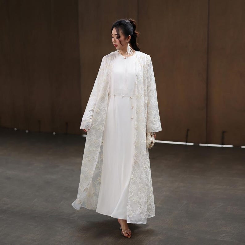 May include: A woman wearing a white dress and a long, sheer, embroidered coat. The coat features intricate floral patterns and covers the dress. She is also carrying a small, white clutch.