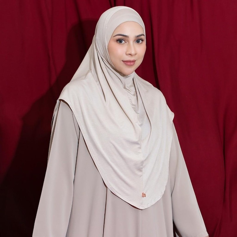 May include: A beige hijab and matching dress are shown against a dark red backdrop. The hijab is draped over the head and shoulders, with a soft, flowing appearance. The dress has long sleeves and a modest neckline, creating a coordinated ensemble.