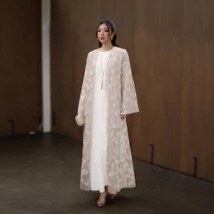May include: A woman wearing a long, white dress with a beige, floral-embroidered overcoat. The overcoat has wide sleeves and a flowing design. She is also carrying a small, white clutch.