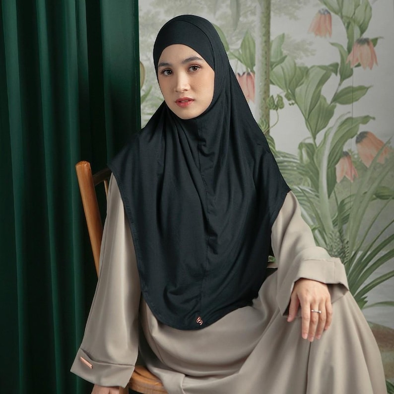 May include: A black hijab with a matching head covering. The hijab is draped over a beige dress. The background features a green curtain and a floral wallpaper. The hijab has a small, copper-colored logo.