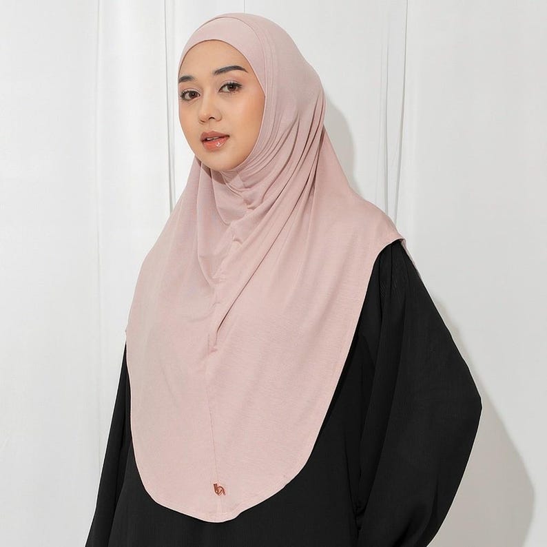 May include: A light pink hijab, a head covering worn by some Muslim women. The hijab drapes over the head and shoulders, with a smooth, soft texture. The person in the image is wearing a black top. The hijab has a small logo on the front.