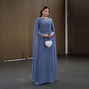 Peut inclure: Longue robe bleu pervenche avec manches longues et cape fluide. La robe présente des ornements délicats sur les manches et le décolleté. Une pochette blanche en forme de cœur est tenue devant.