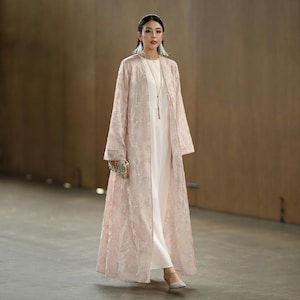 May include: A full-length, pale pink embroidered duster coat over a white dress. The coat features a delicate floral pattern and long sleeves. The woman is holding a small, embellished clutch and wearing a long necklace and earrings. The outfit is completed with matching shoes.