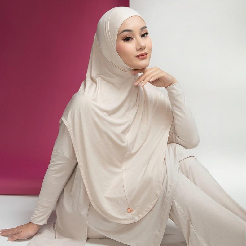 May include: A beige hijab and matching long-sleeved top and pants set. The hijab drapes over the head and shoulders, and the outfit is made of a soft, flowing fabric. The model is sitting with a pink and white background.