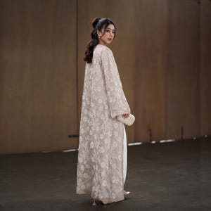 May include: A long, beige embroidered coat with a floral pattern. The coat is worn over a white outfit and paired with a small, white beaded clutch. The sleeves are wide and the coat reaches the floor. The person is wearing silver heels.