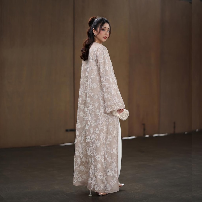May include: A long, beige embroidered coat with a floral pattern. The coat is worn over a white outfit and paired with a small, white beaded clutch. The sleeves are wide and the coat reaches the floor. The person is wearing silver heels.