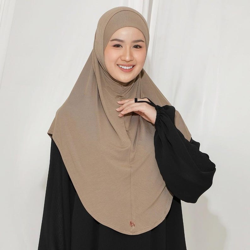 May include: A tan hijab with a matching head covering. The hijab is draped over the shoulders and chest. The person is wearing a black long-sleeved top. The background is a plain white.