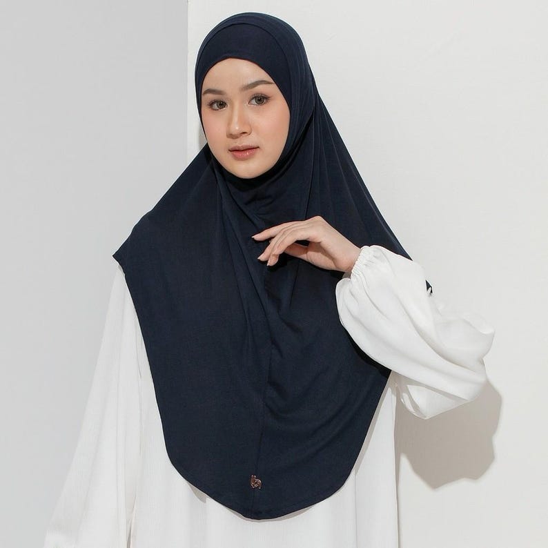 May include: A navy blue hijab, a head covering, is draped over the head and shoulders. The hijab is made of a soft, flowing fabric. The person is wearing a white blouse with puffy sleeves. A small, decorative emblem is visible on the hijab.