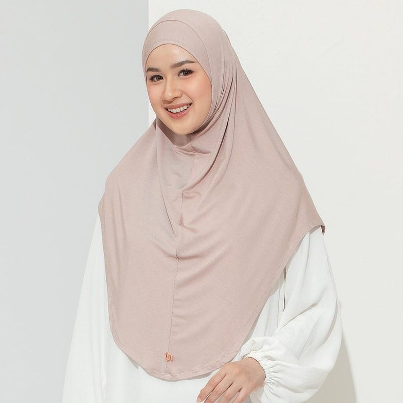 May include: A light brown hijab, a head covering worn by some Muslim women. The hijab is draped over the head and shoulders. The person in the image is wearing a white blouse. The background is a plain white wall.