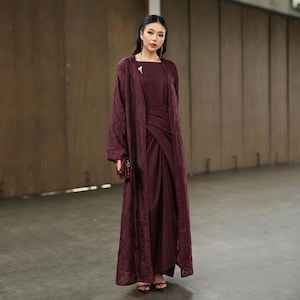 May include: A burgundy outfit comprising a long, flowing overcoat with a textured pattern, worn over a matching dress. The dress has a draped design. A small, brown clutch and sandals complete the look. A small brooch is pinned on the overcoat.