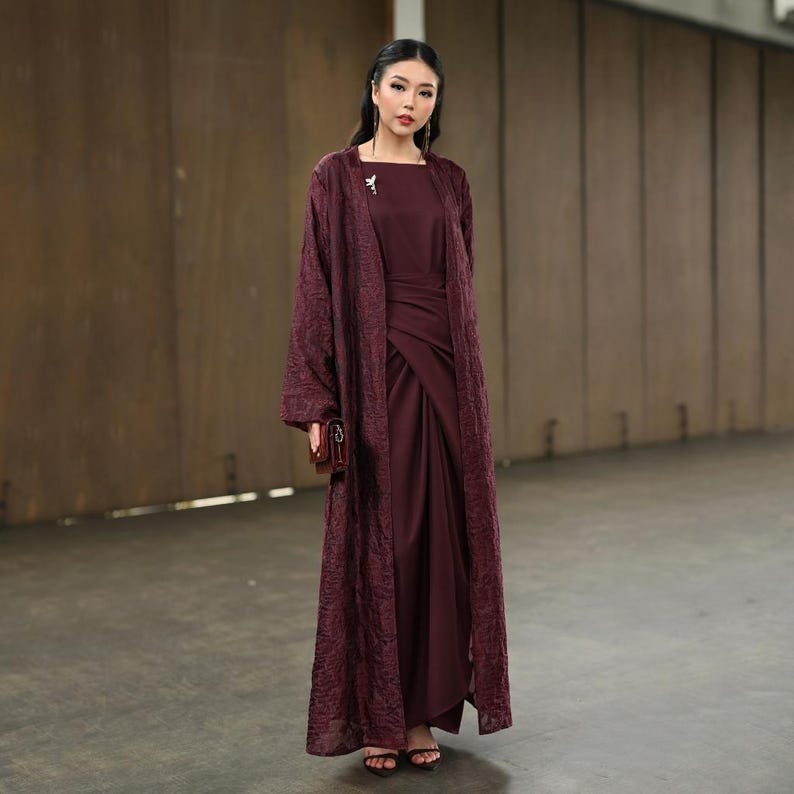 May include: A burgundy outfit comprising a long, flowing overcoat with a textured pattern, worn over a matching dress. The dress has a draped design. A small, brown clutch and sandals complete the look. A small brooch is pinned on the overcoat.