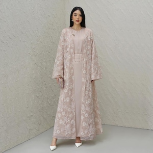 May include: A blush-colored formal dress ensemble. The outfit includes a long, flowing dress, a matching embroidered overcoat, and a small clutch. The overcoat features intricate white floral embroidery. The model is wearing white heels.