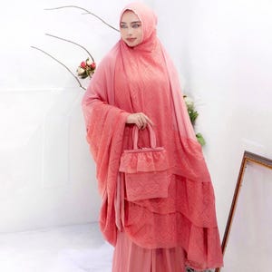 May include: A coral-colored prayer outfit, including a head covering, layered dress, and matching handbag. The outfit features a delicate lace pattern and a soft, flowing design. The ensemble is a modest and elegant choice for prayer.