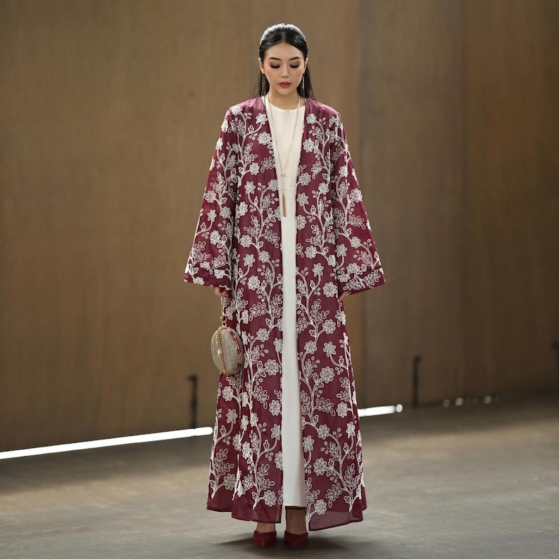 May include: A long, burgundy open-front robe with white floral embroidery. The robe is worn over a white outfit and paired with red shoes. A small, round purse is held in the hand.