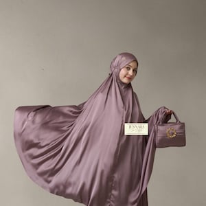 May include: A mauve satin prayer garment featuring a matching head covering and a small handbag with a gold circular embellishment. The fabric has a smooth, reflective finish. A rectangular label with the text JENNARA is visible on the garment.