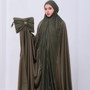 May include: An olive green prayer garment set. The set includes a head covering, a long dress, and a matching bag. The head covering and the center of the dress feature a delicate lace pattern. The bag has a gold-coloured clasp.