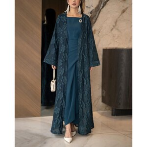 May include: A teal-coloured, floor-length dress with a matching floral lace overcoat. The dress has a square neckline and a draped skirt. A small, pearl-handled purse and white heels complete the ensemble. A small brooch is pinned on the dress.