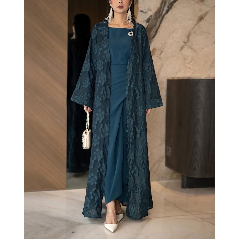 May include: A teal-coloured, floor-length dress with a matching floral lace overcoat. The dress has a square neckline and a draped skirt. A small, pearl-handled purse and white heels complete the ensemble. A small brooch is pinned on the dress.