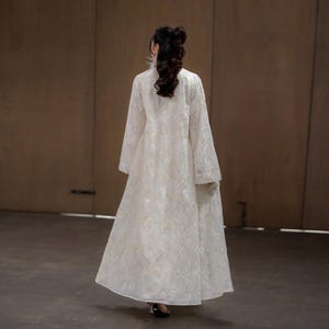 May include: A long, white, embroidered coat with a high collar and long sleeves. The coat features a delicate floral pattern and a flowing silhouette. The garment is worn with black heels and a small, embellished clutch.