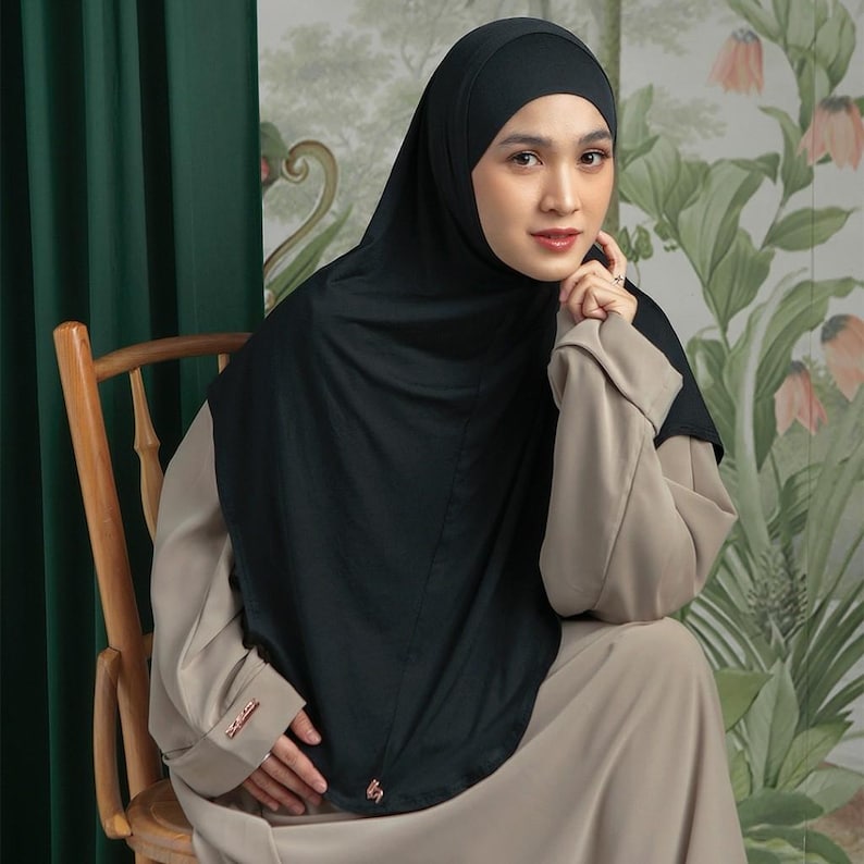 May include: A black hijab and matching head covering. The woman is wearing a beige dress and sitting on a wooden chair. The background features a floral design and a green curtain.