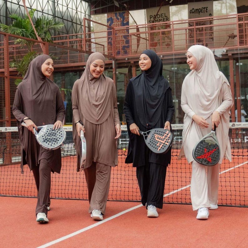 May include: Four women wearing athletic hijabs and matching outfits, each holding a padel racket, stand on a red court. The outfits are in shades of brown, black, and beige. The background includes a tennis net and signs.