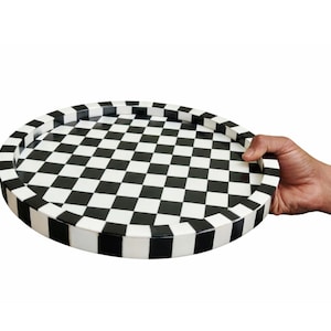 May include: A round decorative tray with a black and white checkered pattern. The tray has a raised edge and is held by a hand. The tray's design is reminiscent of a chessboard.
