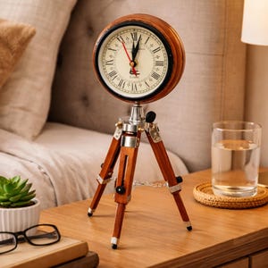 May include: A vintage-style clock with a round wooden frame and a white face with black Roman numerals. The clock sits atop a silver and wood tripod stand. The clock's hands are red. The clock is on a wooden nightstand.