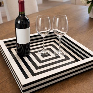 Puede incluir: Una bandeja cuadrada con un patrón geométrico en blanco y negro contiene una botella de vino tinto y dos copas de vino. La botella de vino tiene una etiqueta blanca en blanco. La bandeja está sobre una superficie de madera.