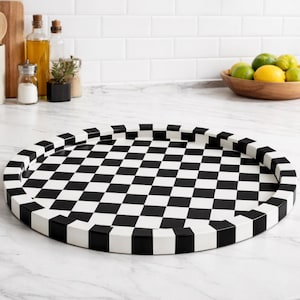 Puede incluir: Una bandeja redonda para servir con un patrón de cuadros blancos y negros. La bandeja tiene un borde elevado con bloques alternos blancos y negros. Está sobre una encimera blanca en una cocina.