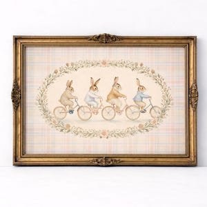 May include: A framed wall art print featuring four rabbits riding bicycles in a line, enclosed within an oval floral wreath. The background has a soft plaid pattern in pastel pink, blue, and cream. The frame is ornate, metallic gold, and rectangular.