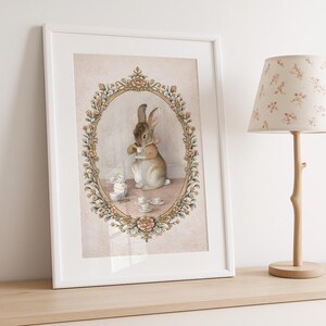 May include: A framed print featuring a watercolor illustration of a rabbit at a tea party. The rabbit, in shades of brown and beige, is adorned with a pink bow. The artwork is set within an ornate, oval-shaped frame with floral accents, and includes miniature teacups.