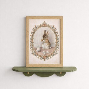 May include: A framed print featuring a watercolor illustration of a rabbit enjoying tea. The artwork is set within an oval floral frame, with a light brown wooden frame. The rabbit is depicted with a pink bow, surrounded by teacups and a small mouse.