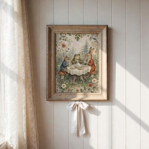 May include: A framed print depicts a whimsical tea party scene with a mouse, frog, and rabbit seated around a table. The artwork features soft colors, a rustic wooden frame, and a floral and forest backdrop. A cream-colored bow hangs below the frame.
