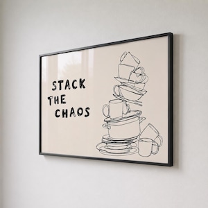 May include: A framed wall art print featuring a black line drawing of a precarious stack of kitchenware including mugs, bowls, and a pot on a light beige background. The text STACK THE CHAOS is printed in black on the left side of the artwork.