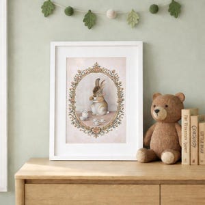 May include: A framed print featuring a watercolor illustration of a rabbit enjoying tea. The artwork is set within an oval floral frame, with a soft pink background. A stuffed teddy bear and children's books are on a wooden surface.