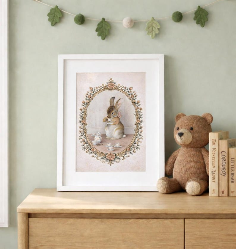 May include: A framed print featuring a watercolor illustration of a rabbit enjoying tea. The artwork is set within an oval floral frame, with a soft pink background. A stuffed teddy bear and children's books are on a wooden surface.