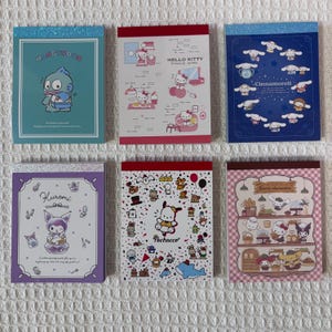 May include: Six colorful memo pads featuring popular cartoon characters. Each pad has a different design, including Hello Kitty, Cinnamoroll, Kuromi, and Pochacco. The pads have glittery borders and are arranged on a white surface.